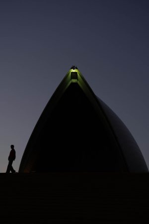 Silhouette of a person walking beneath a Sydney Opera House sail at blue hour, with a light glowing at the sail's tip