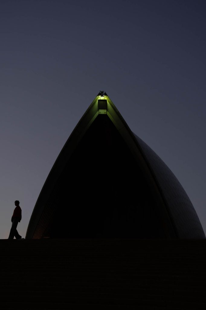 Silhouette of a person walking beneath a Sydney Opera House sail at blue hour, with a light glowing at the sail's tip