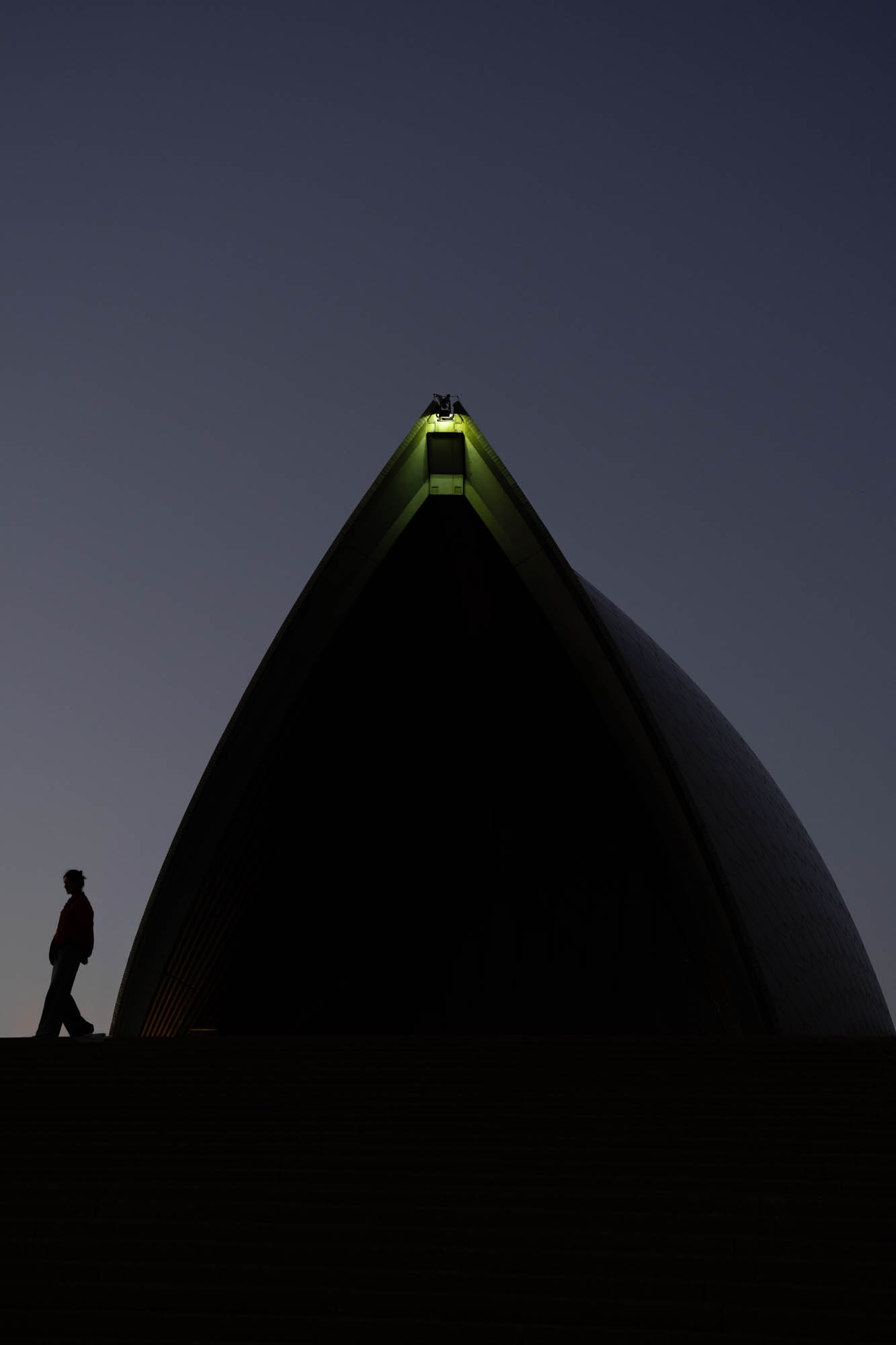 Beneath the Sail Silhouette of a person walking beneath a Sydney Opera House sail at blue hour, with a light glowing at the sail's tip
