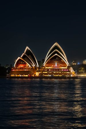 Sydney Opera House lit up at night with glowing sails and golden foyers, reflected in the dark water of Sydney Harbour