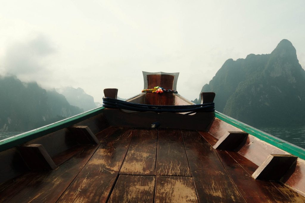 Bow of a wooden longtail boat heading towards misty limestone cliffs in Khao Sok National Park, Thailand