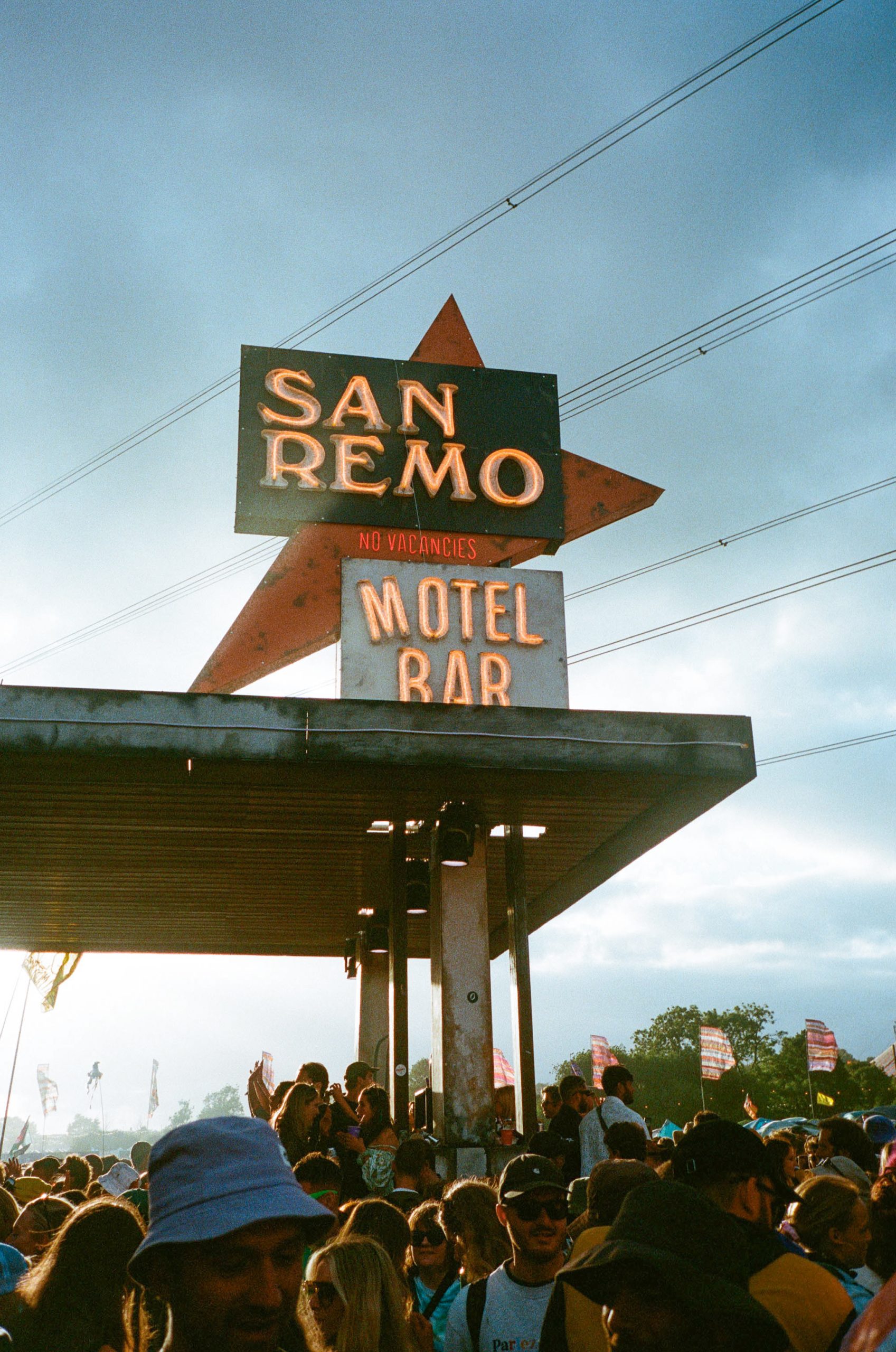 San Remo, Glastonbury Neon San Remo Motel Bar sign with a red star and No Vacancies light above a packed festival crowd at Glastonbury, under a blue evening sky